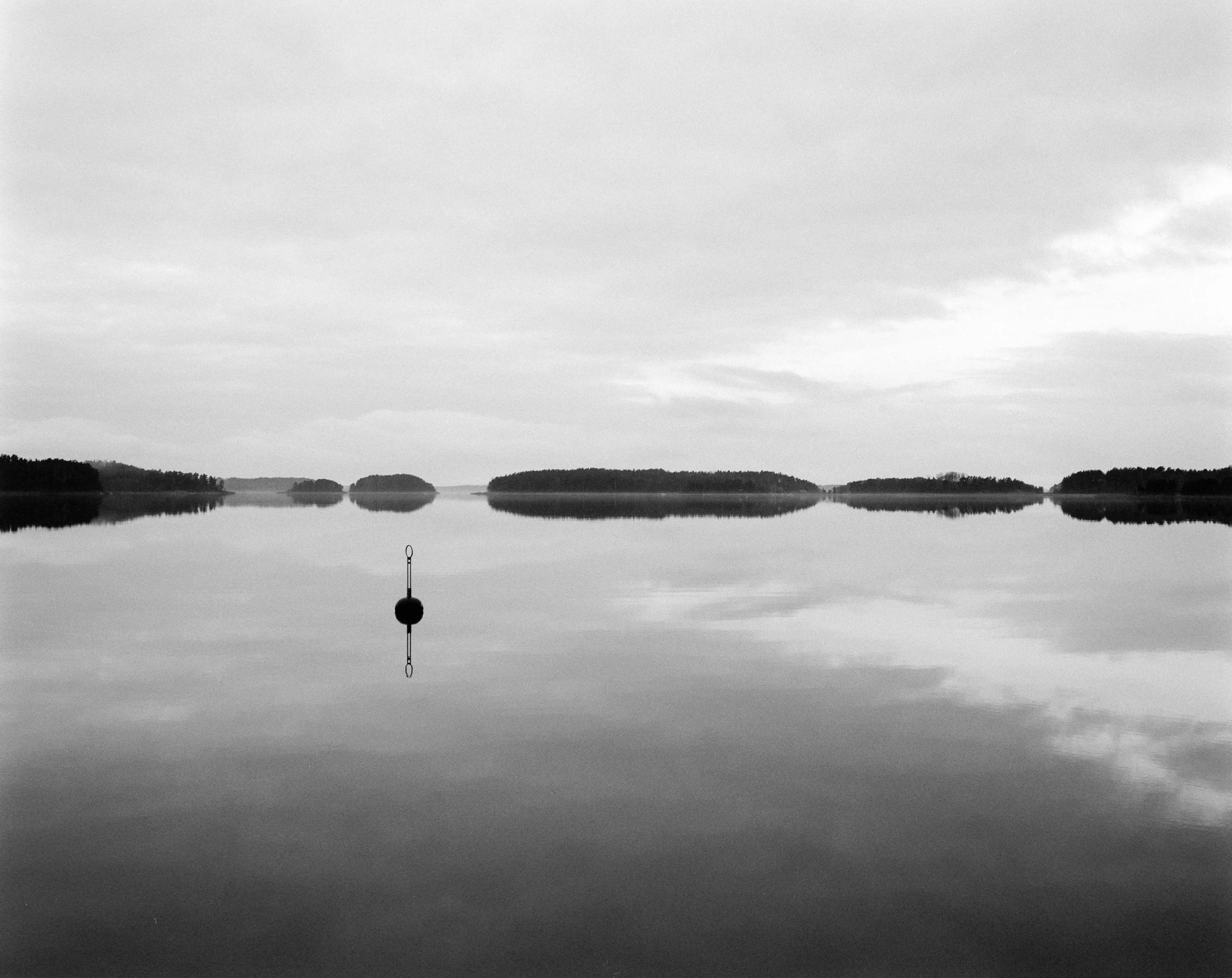 A solitary buoy on a mirror-still lake under an overcast sky, wooded islands on the horizon