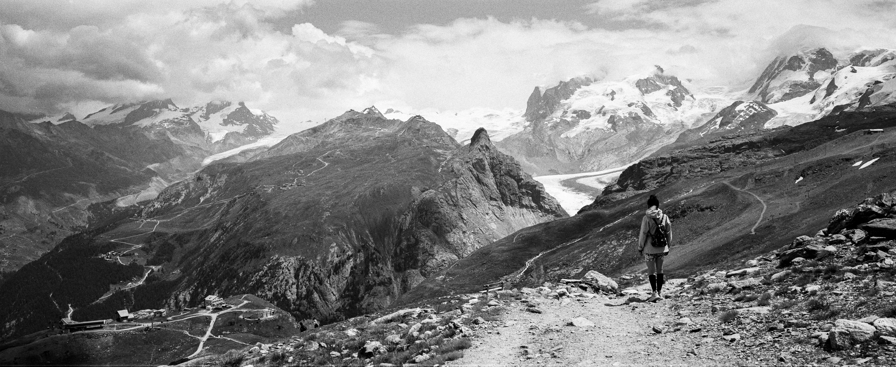 A hiker on a rocky alpine trail before a sweep of glaciated peaks, in black and white