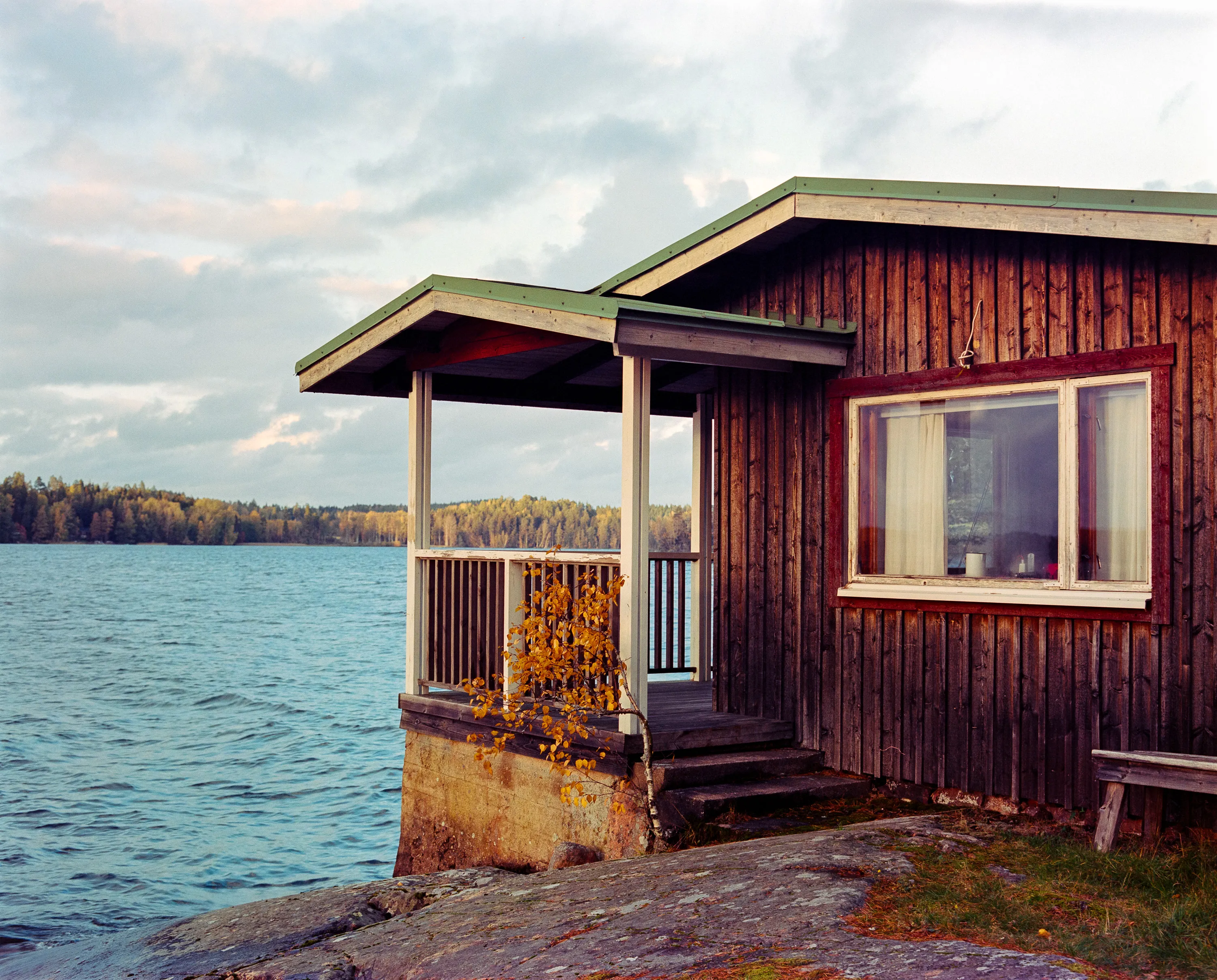 A wooden sauna cabin on lakeshore rocks, a small birch in autumn color by its steps