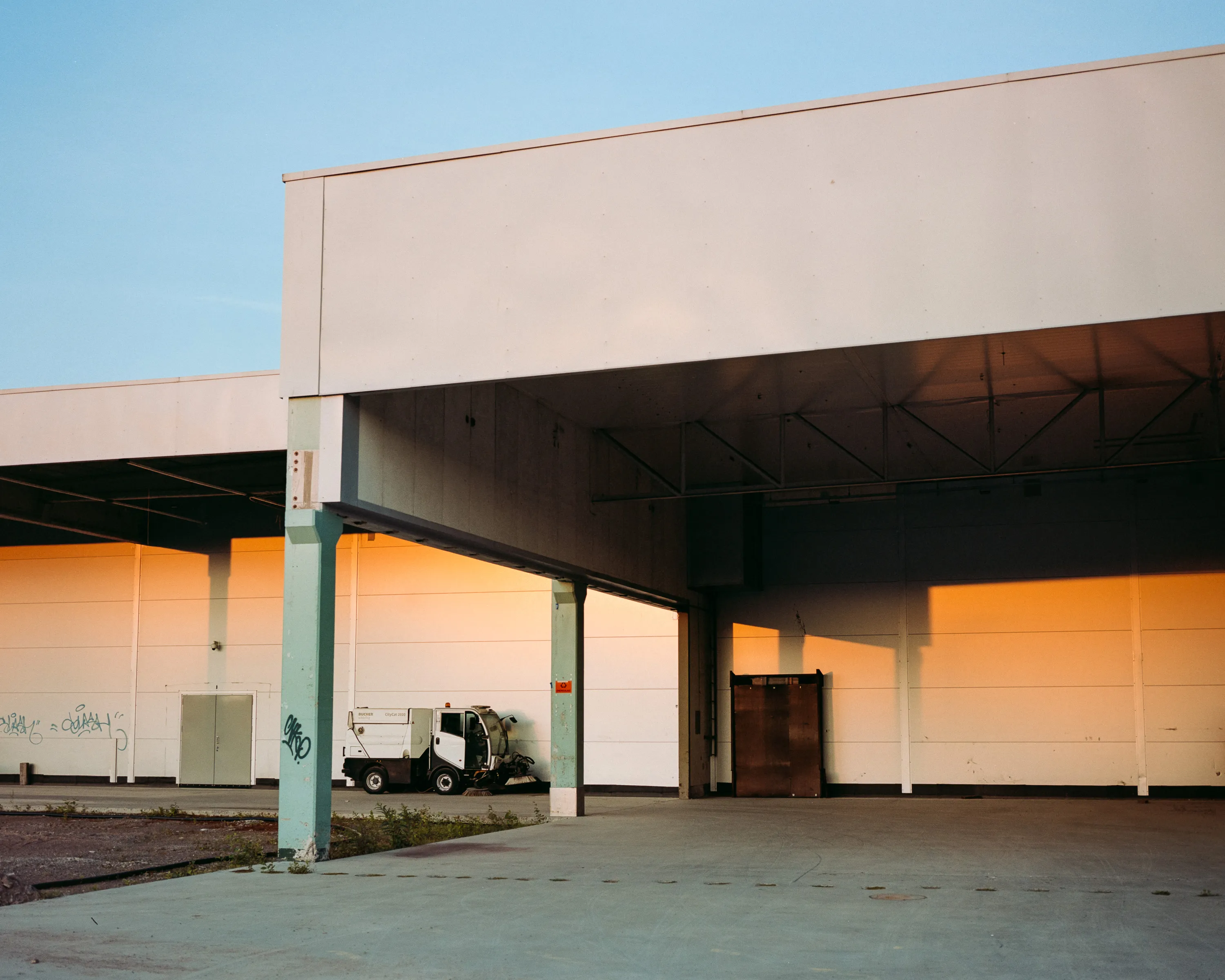 An empty warehouse loading bay at sunset, low orange light across white walls, a street sweeper parked