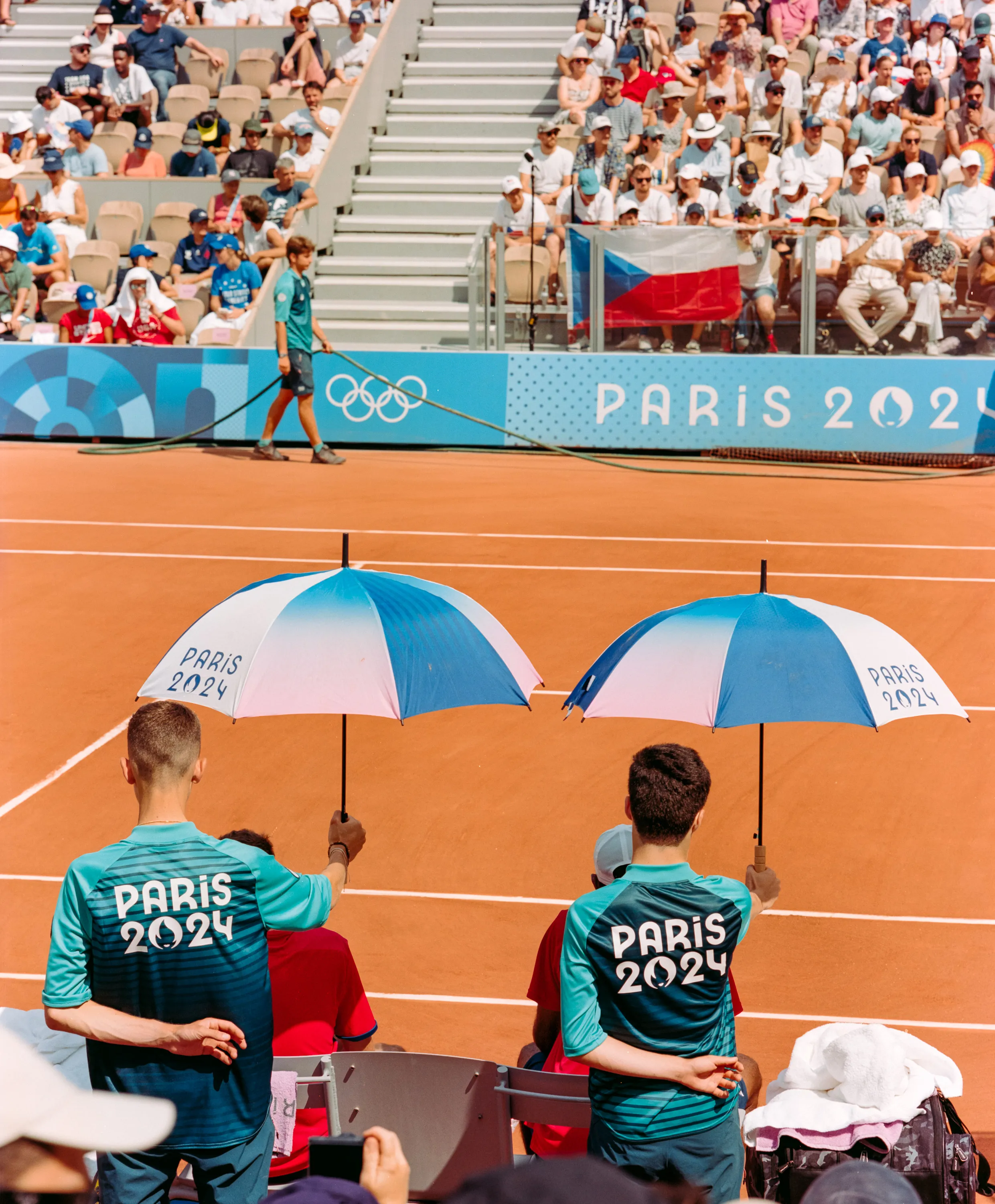 Two ball kids holding Paris 2024 umbrellas on a clay tennis court